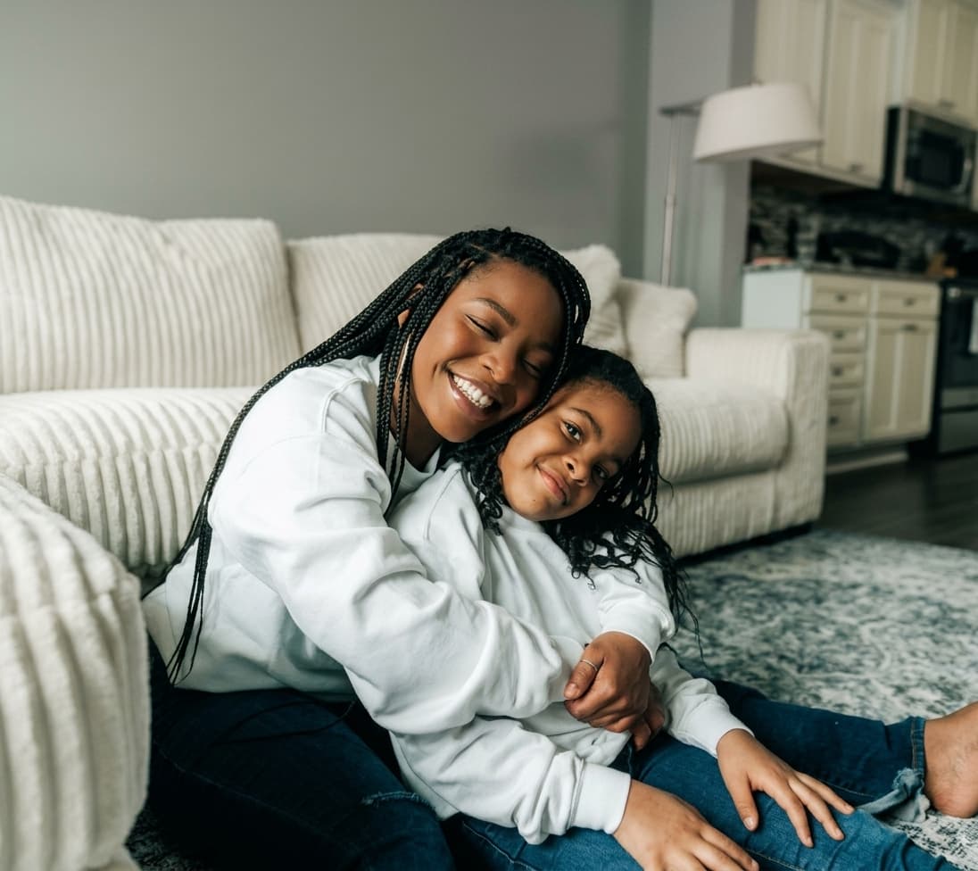 A smiling mother hugging her daughter as they sit together on the living room floor