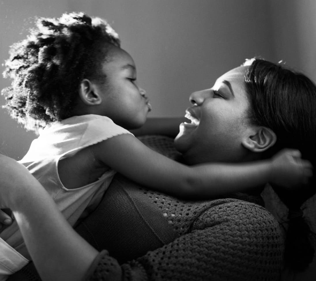 A laughing mother holding her young daughter, who leans in for a kiss